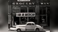 A large sign reading "I am an American" hangs in the window of a shuttered Oakland grocery store the day after Pearl Harbor, as a 1940s car sits in the foreground.