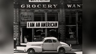 A large sign reading "I am an American" hangs in the window of a shuttered Oakland grocery store the day after Pearl Harbor, as a 1940s car sits in the foreground.