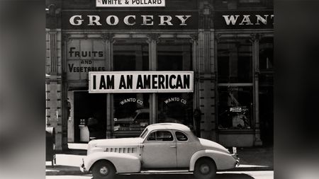 A large sign reading "I am an American" hangs in the window of a shuttered Oakland grocery store the day after Pearl Harbor, as a 1940s car sits in the foreground.