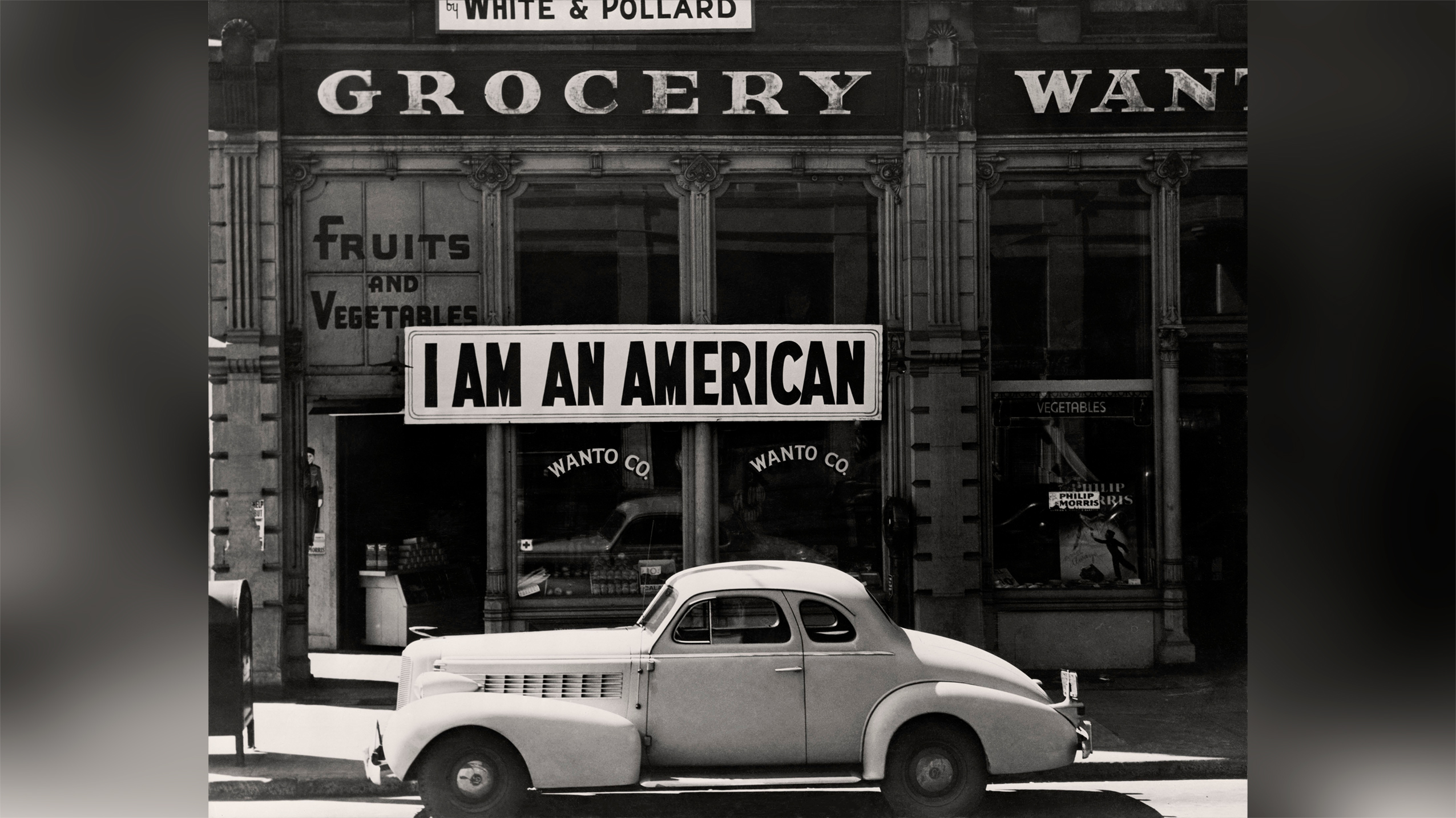 A large sign reading "I am an American" hangs in the window of a shuttered Oakland grocery store the day after Pearl Harbor, as a 1940s car sits in the foreground.