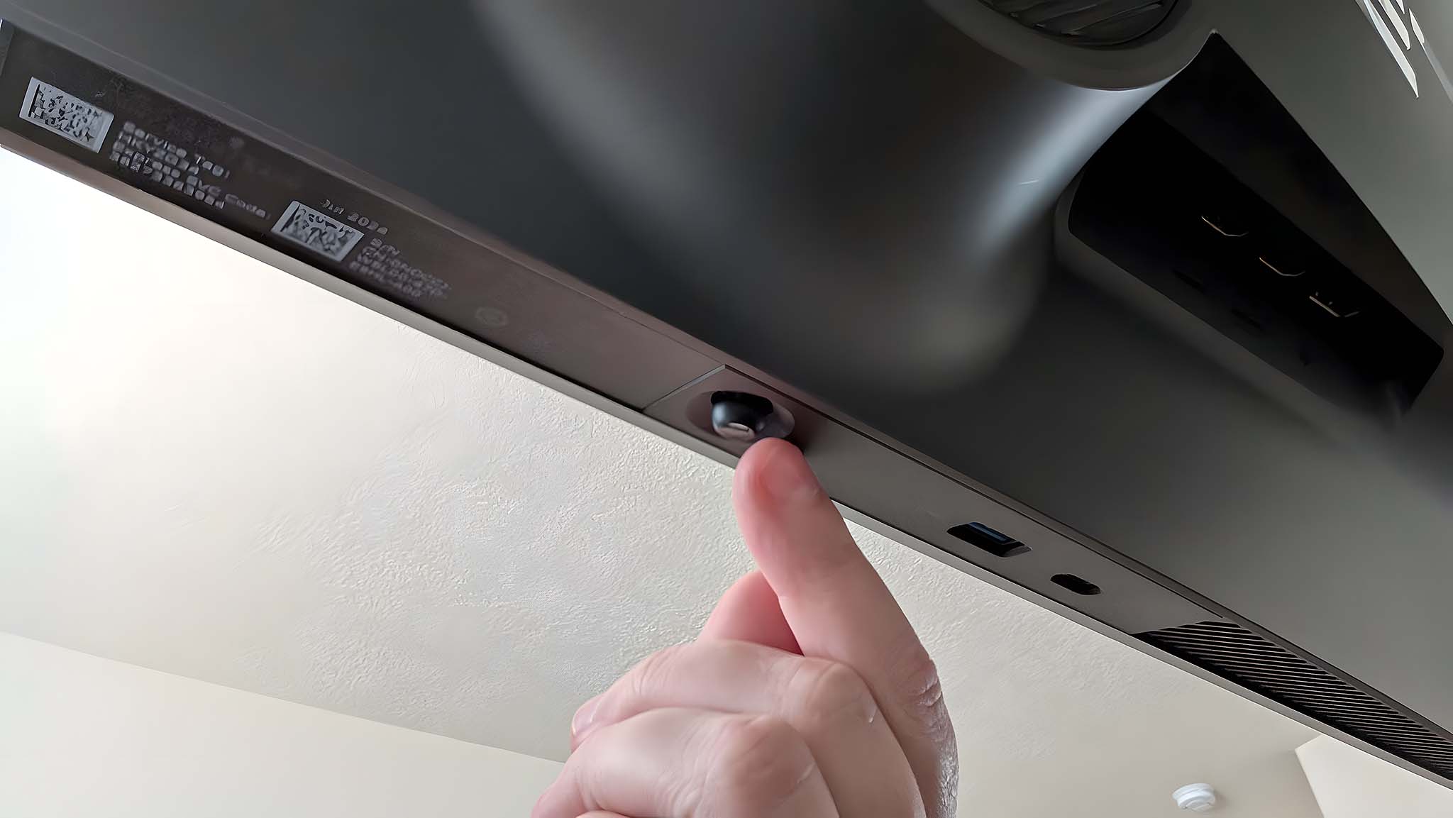 A hand reaching under the monitor to interact with the Alienware AW2725QF Gaming Monitor's OSD controls.