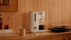 A white crop coffee maker with a glass carafe on a wooden sideboard beside a record player. 