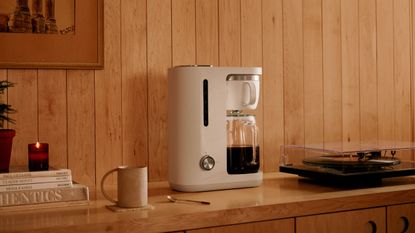 A white crop coffee maker with a glass carafe on a wooden sideboard beside a record player. 