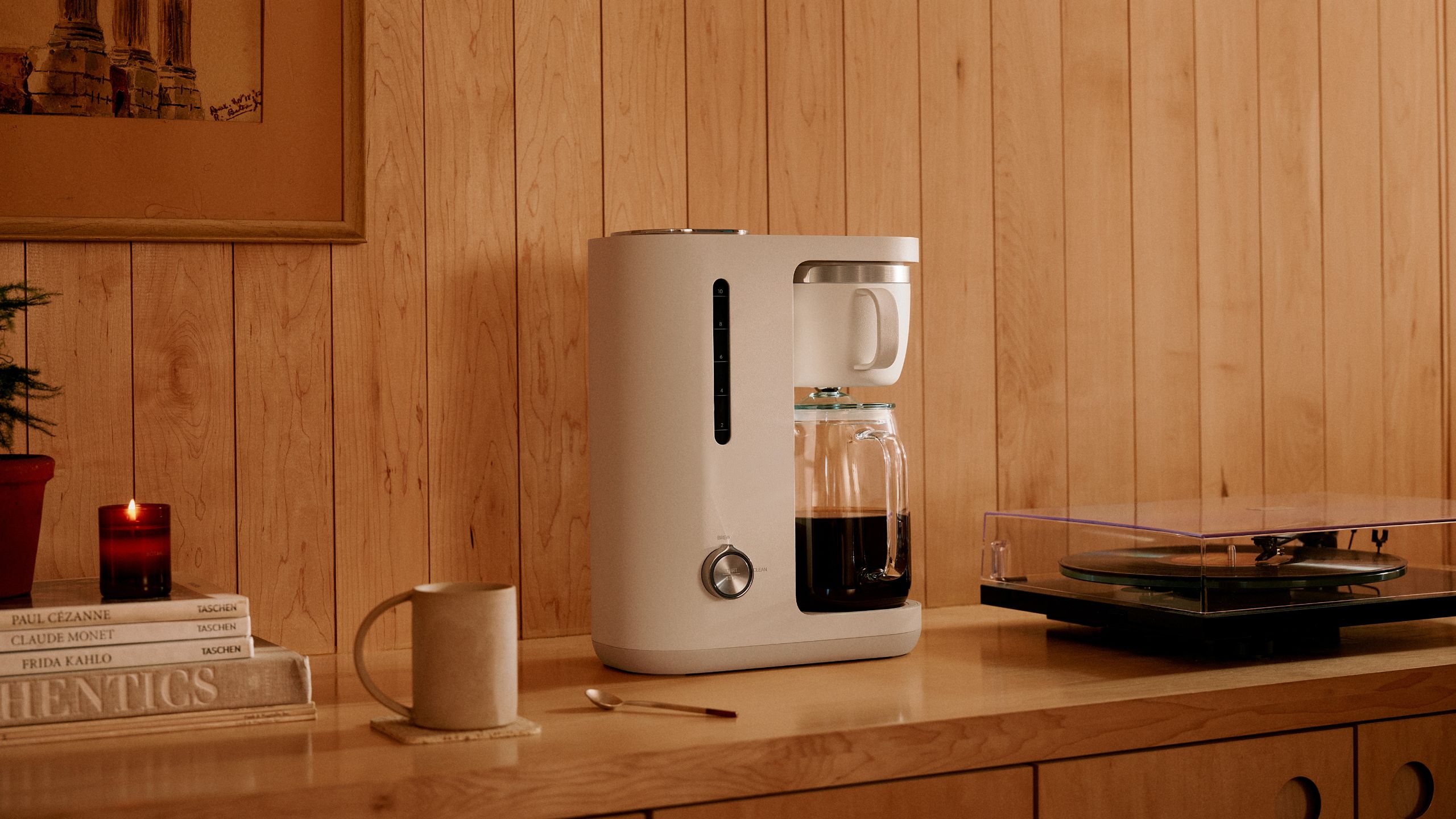 A white crop coffee maker with a glass carafe on a wooden sideboard beside a record player. 