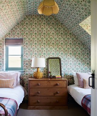 twin attic cottage room with morris & co wallpaper over walls and ceiling, metal beds and wooden chest of drawers