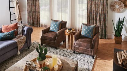 Living room with natural, earth-toned colors, with two brown armchairs, a large gray sofa, a white patterned rug, floor-to-ceiling curtains, and a coffee table with plants and candles, on wooden floor.