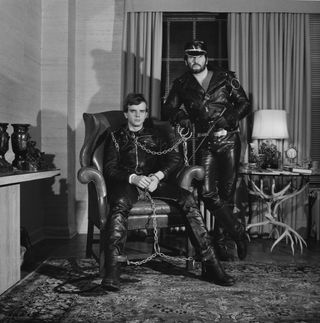 Two males wearing black leather outfits and chained to each other