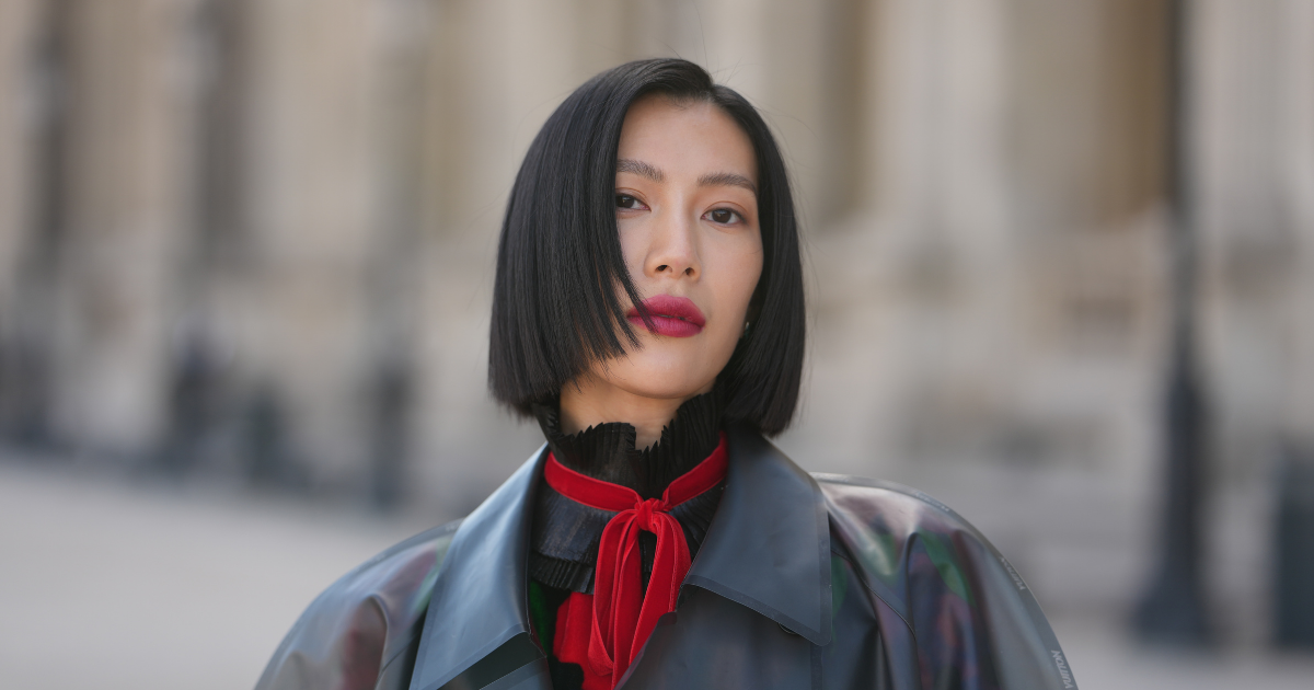 Hair Longevity A guest wears a straight chin-length black bob with a deep side part, a black pleated ruffled high-neck top, a red velvet scarf tied in a bow at the neck, a dark grey iridescent vinyl coat with a wide collar and glossy finish, outside Louis Vuitton, during Paris Fashion Week