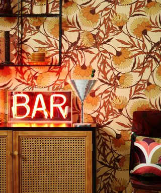 A wall with retro orange leaves wallpaper, a rattan wooden console table with a red neon sign that says 'bar' and a dark brown chair with a retro pillow next to it