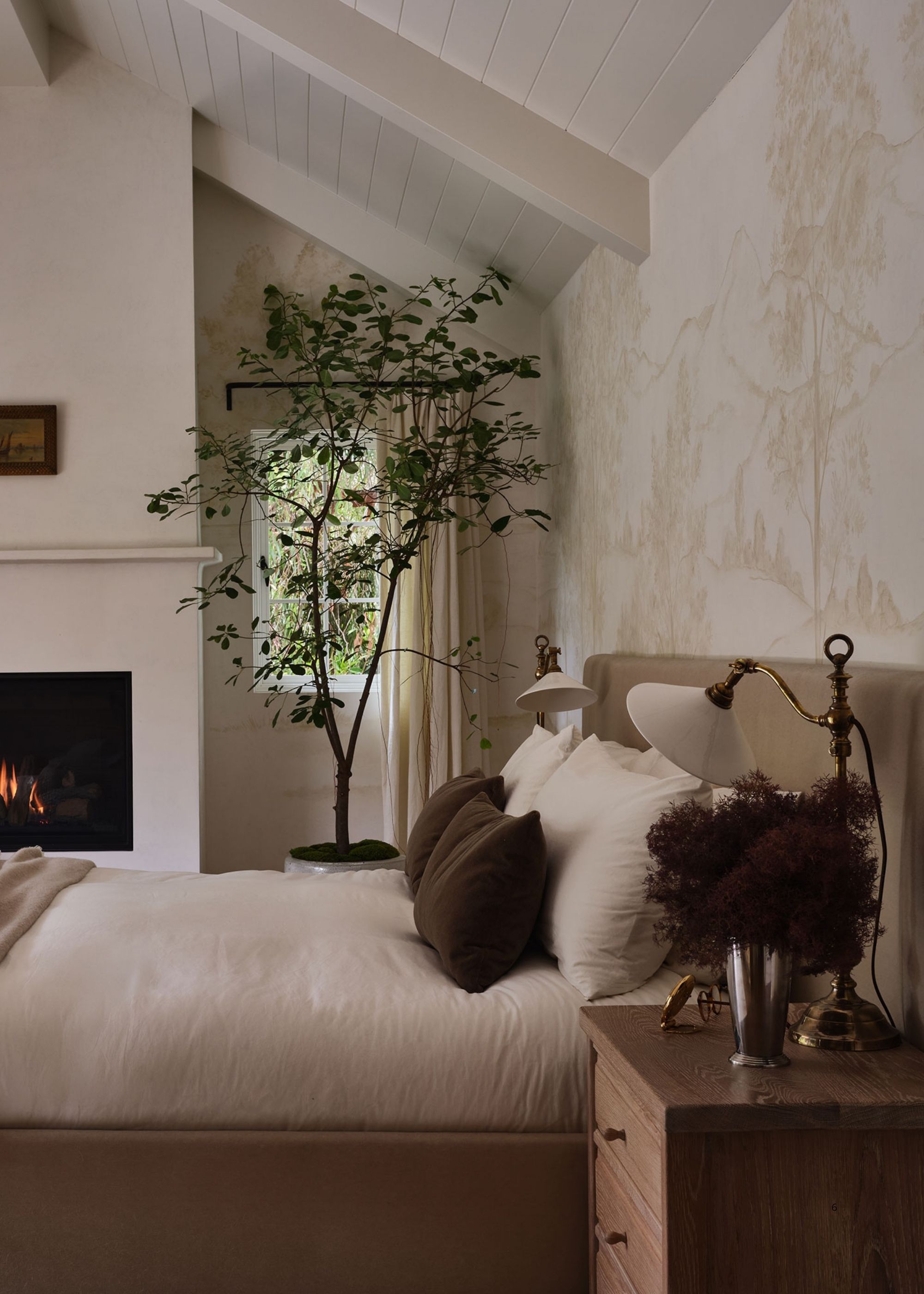 Image of a modern, neutral bedroom with a tan velvet bed that has white bedding and dark brown throw pillows. There is a tree in the corner by a fireplace.