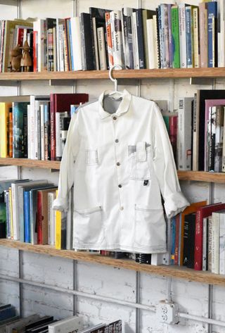 A white workwear jacket with buttons hangs from a wooden shelving unit filled with colorful books in a studio setting.