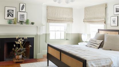 bedroom blind ideas, green and white bedroom with sage green half painted walls, rattan and wood bed, fireplace, oatmeal checked Roman blinds, artwork, vases