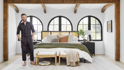 bobby berk in a bedroom with white walls, wood beams, and bed with rattan headboard