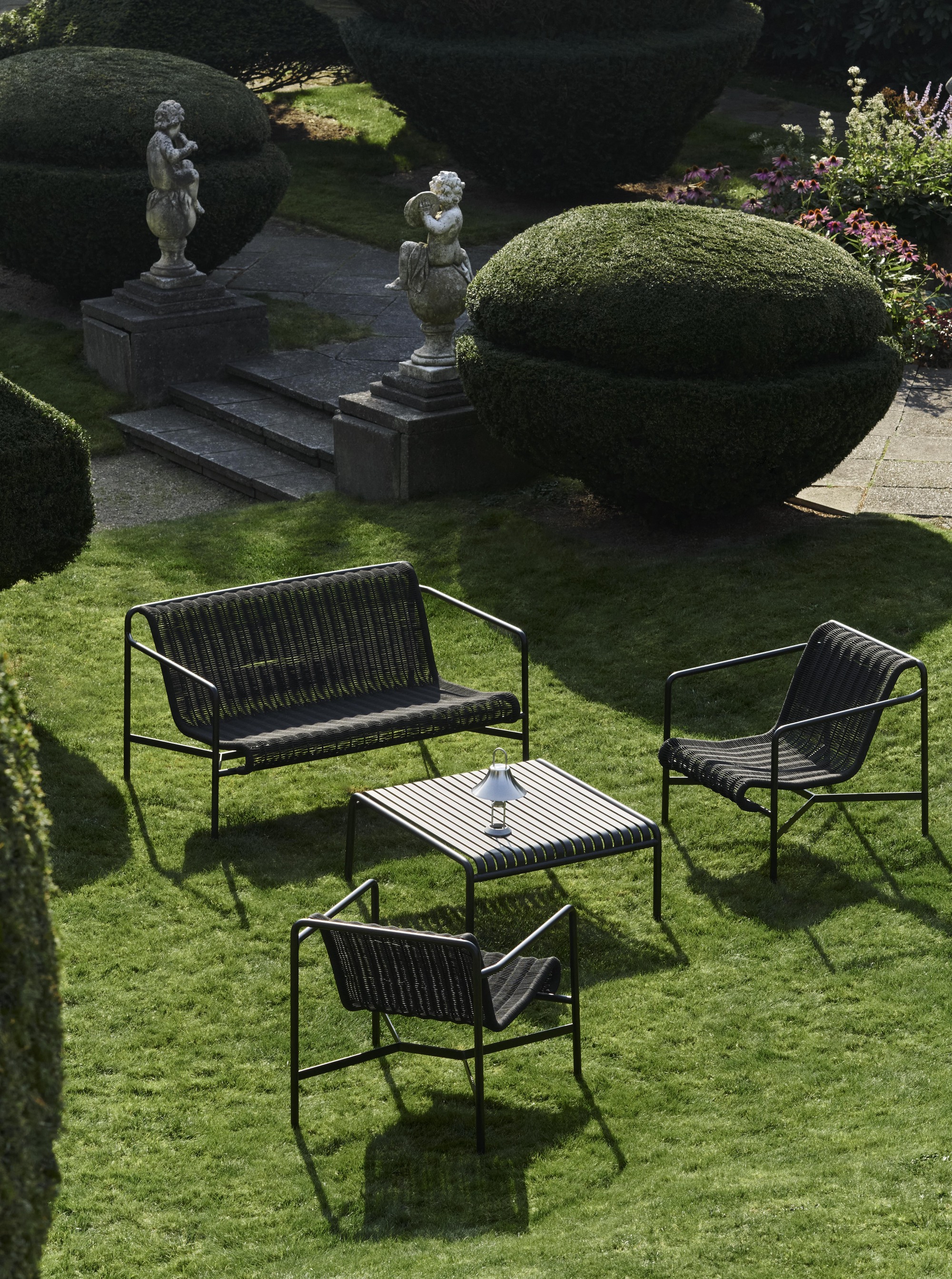Image of an outdoor black rope bench styled by two matching armchairs and a black metal table. They are on a grassy lawn with manicured shrubs and stone statues.