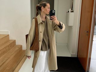 Dutch fashion influencer Anouk Yve wearing an oversize trench coat, a tan sweater, white linen pants, and suede shoulder bag