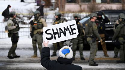 An onlooker holds a sign that reads "Shame" as members of law enforcement work the scene following a suspected shooting by an ICE agent during federal law enforcement operations