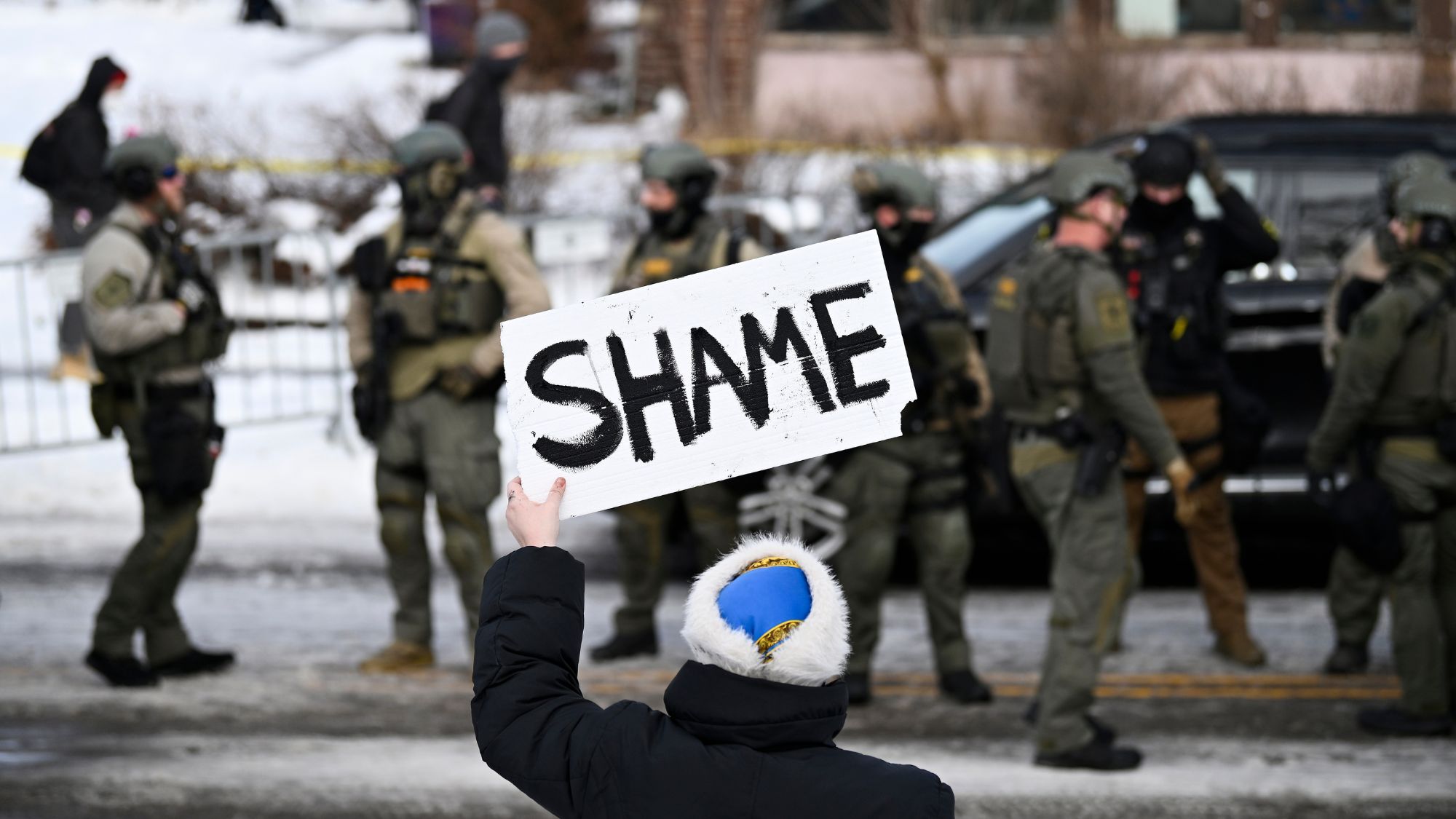 An onlooker holds a sign that reads "Shame" as members of law enforcement work the scene following a suspected shooting by an ICE agent during federal law enforcement operations