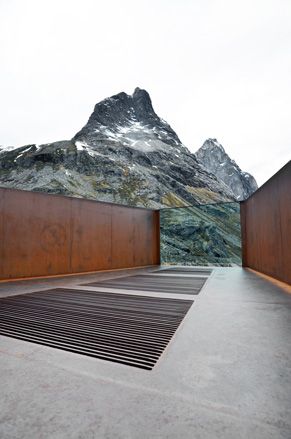 An open top viewing platform with concrete floors, rusted steel walls and a glass panel. A mountain can also be seen under a clear white sky