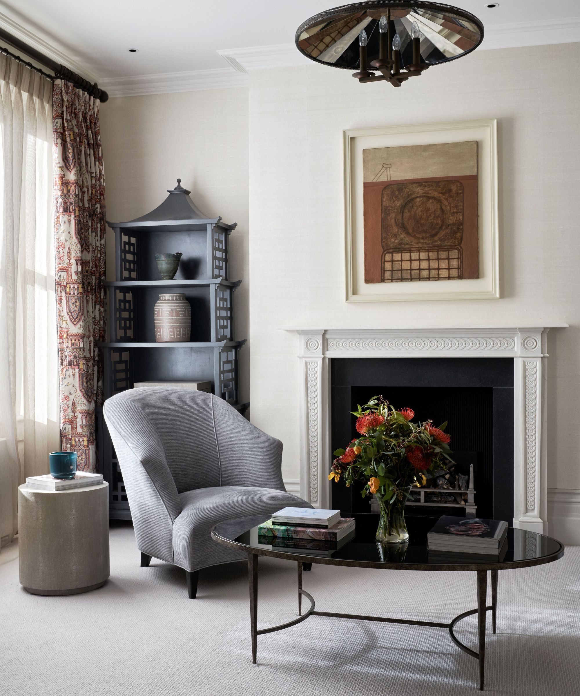Living room with layered curtains, grey armchair, black lacquer coffee table, fireplace with art above, and antique bookcase with vases