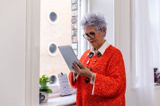 A medium shot of a stylish older woman with silver hair and a bright red sweater is on a tablet by a window, exuding confidence and warmth. Perfect for themes of senior professionals and workplace positivity.