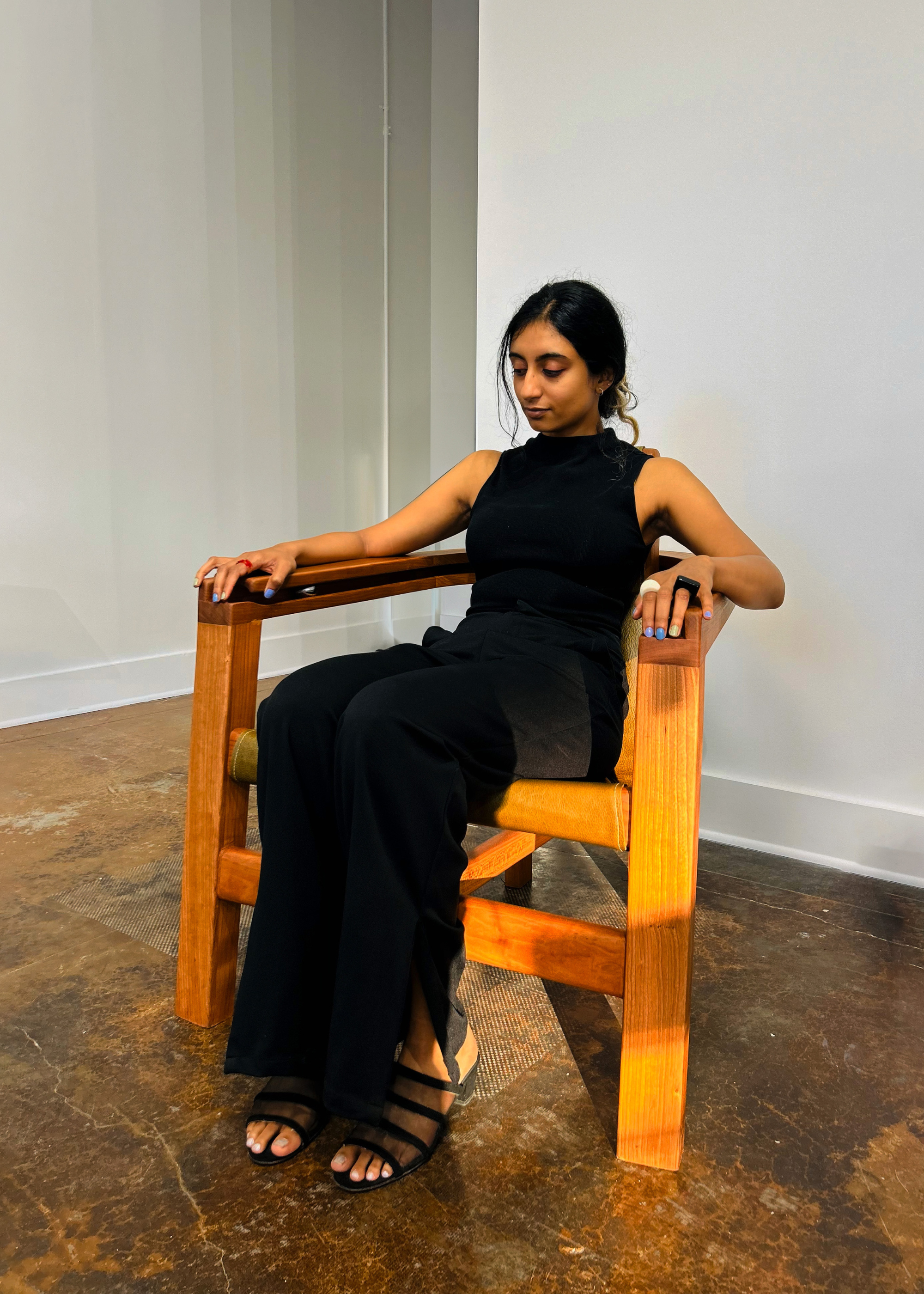 A girl in a black pantsuit sitting in a cherry wood chair in a white room with distressed flooring