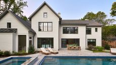 A large, two-story white brick house with black window frames, featuring a stone patio and a luxurious blue swimming pool with an integrated hot tub.