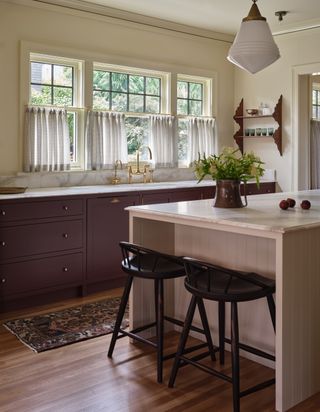 A bright and natural kitchen with plum purple kitchen cabinets and a kitchen island that is painted in a dusty pink color. The countertops are in a white marble and there are half curtains on the windows