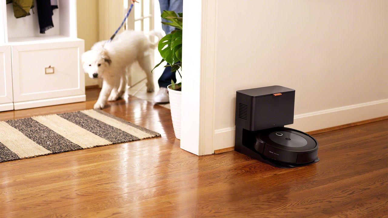 iRobot Roomba j7+ robot vacuum and cleaning base on wooden floor against wall in front of person walking a white dog