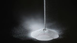 a solid line of a white powdery substance falls from the top, just right of center, into a pile below on a black surface.