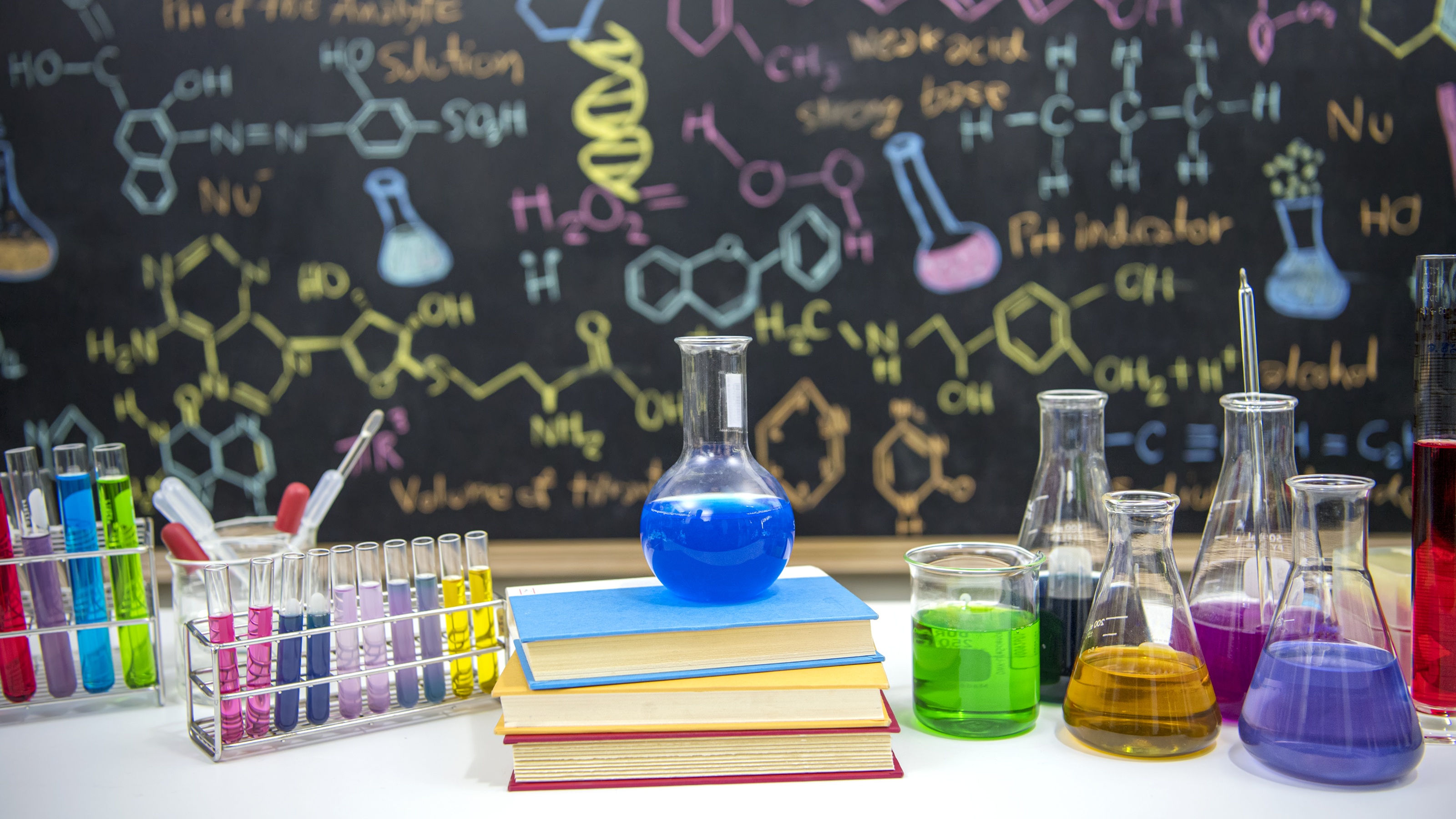 Classroom desk with books and science instruments in front of a chalkboard with a formula drawn in colorful chalk.