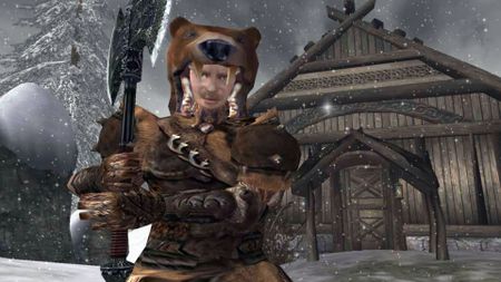 An official screenshot showing a character wearing a bear-pelt outift, holding a large battle axe, standing outside wooden cabin in the snow.