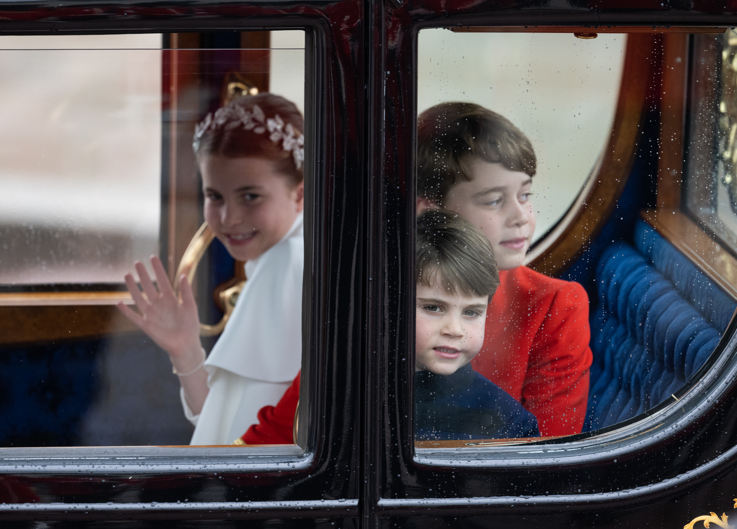 Princess Charlotte wearing a white cape and waving inside of a carriage with Prince Louis and Prince George