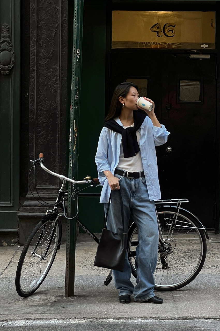 A woman drinking coffee standing on a sidewalk in New York City in front of a bike wearing a navy cardigan tied around the shoulder, a light blue button-down shirt layered over a white tank-top, a black belt, baggy wide-leg jeans, black Tabi ballet flats, and a black tote bag.