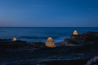 Three woven lanterns photographed on rocks by the sea at dusk