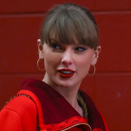 Taylor Swift wearing a red sweater standing in front of a red brick wall and smiling