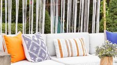 Learn how to create backyard privacy without blocking light. For example, here is a white macrame panel behind a white outdoor seat with orange, purple, and striped throw pillows on it