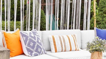 Learn how to create backyard privacy without blocking light. For example, here is a white macrame panel behind a white outdoor seat with orange, purple, and striped throw pillows on it