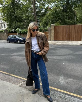 British influencer Lucy Williams poses on a London sidewalk wearing black oversize sunglasses, brown leather jacket, white t-shirt, Khaite studded belt, straight-leg jeans, woven brown tote bag, and The Row black Mary Jane ballet flats