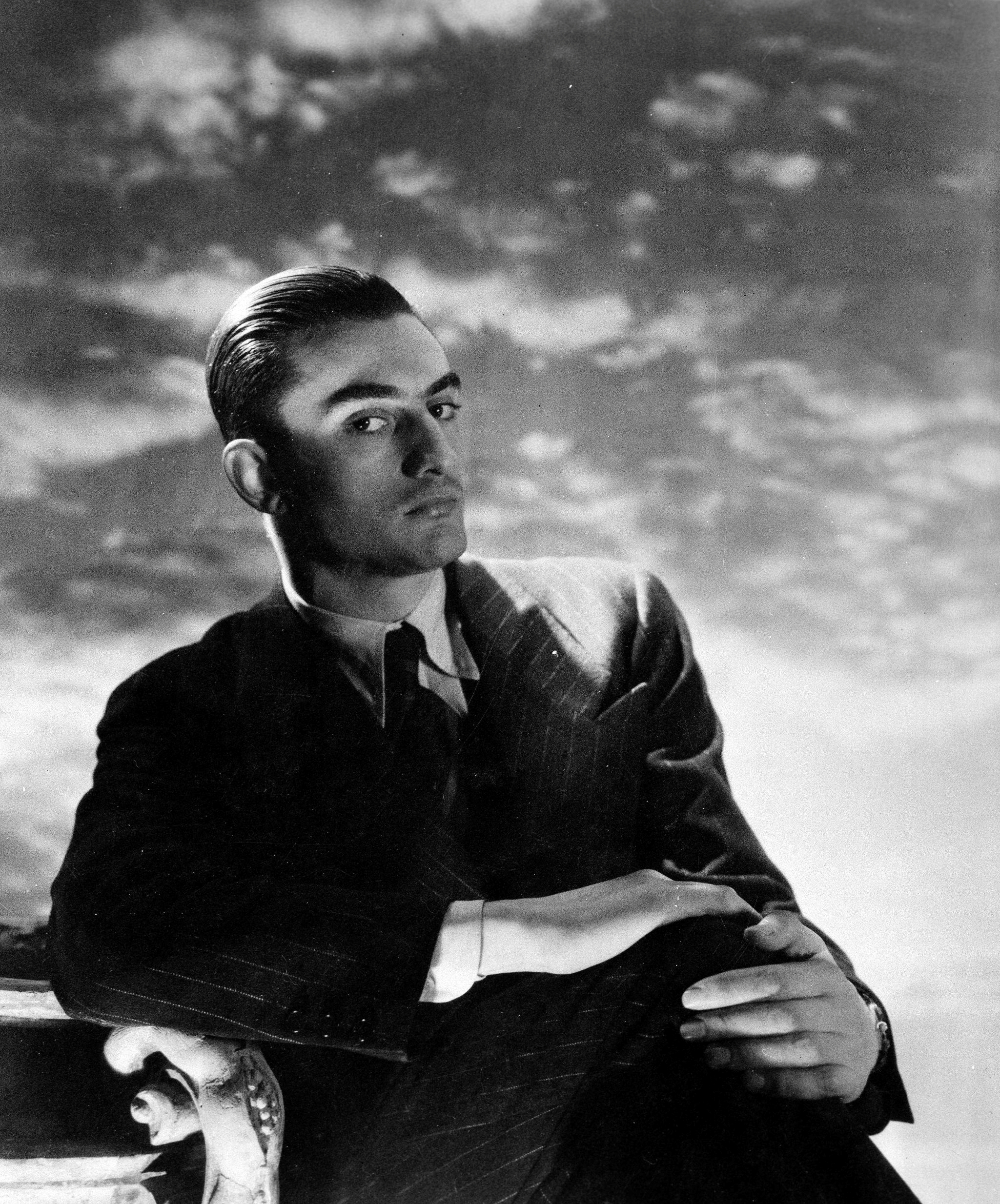 A black-and-white portrait depicts a man in a pinstriped suit sitting with crossed arms against a backdrop of wispy, painted clouds.