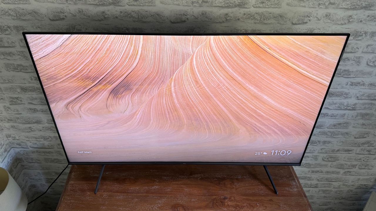 The TCL C6KS 50-inch TV straight-on from above on a TV cabinet in front of a brick wall. On screen is a swirly pink pattern.