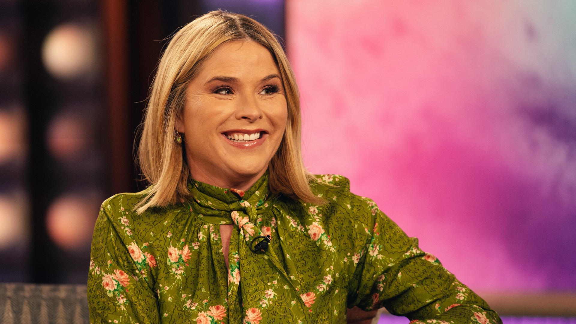 a white woman with short, blonde hair wearing a green, floral print, high neck blouse while being interviewed on the set of a talk show