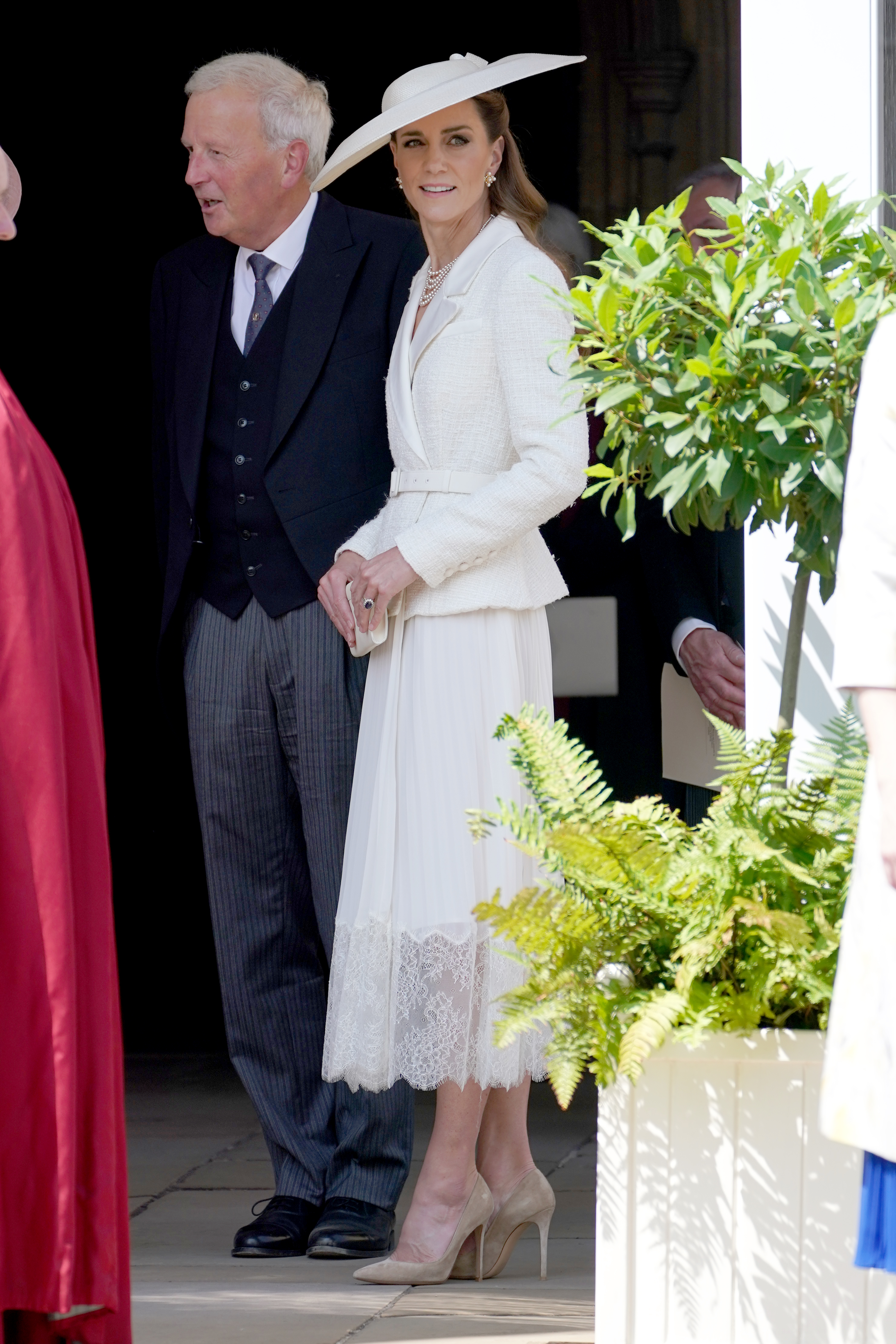 Catherine, Princess of Wales emerges from Windsor Castle wearing a white lace-trim skirt with a tonal jacket neatly fastened around her waist. She wear a wide-brim white hat, dark beige heels and carries a small satin clutch in her hands.