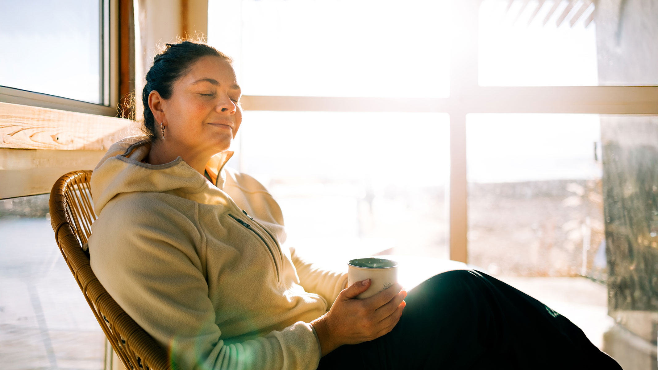 A woman in a hoodie is sitting next to a closed window and relaxing with a cup of tea
