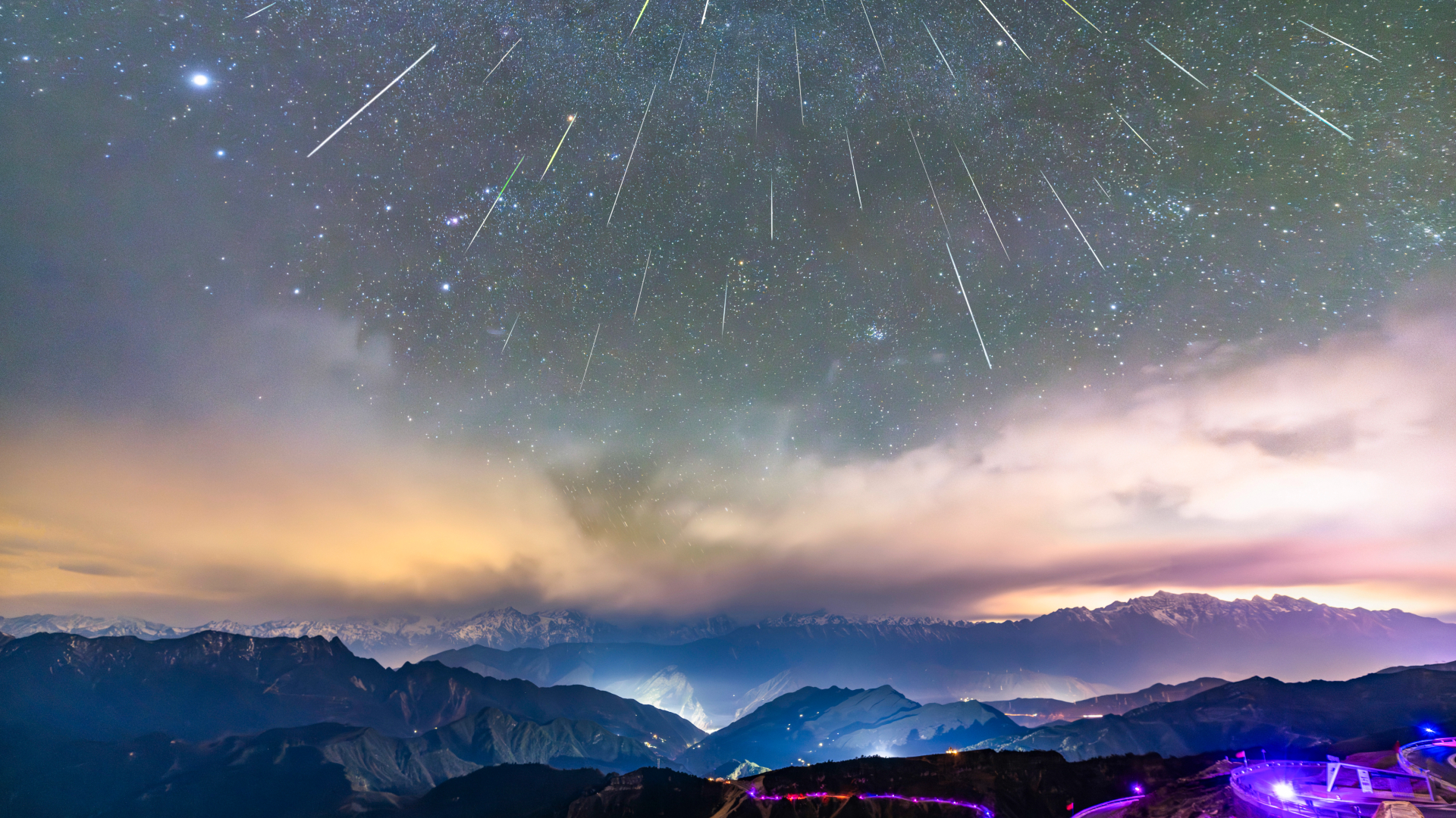 Meteors are pictured swarming downward in a starry sky above a mountain range lit with blue light.