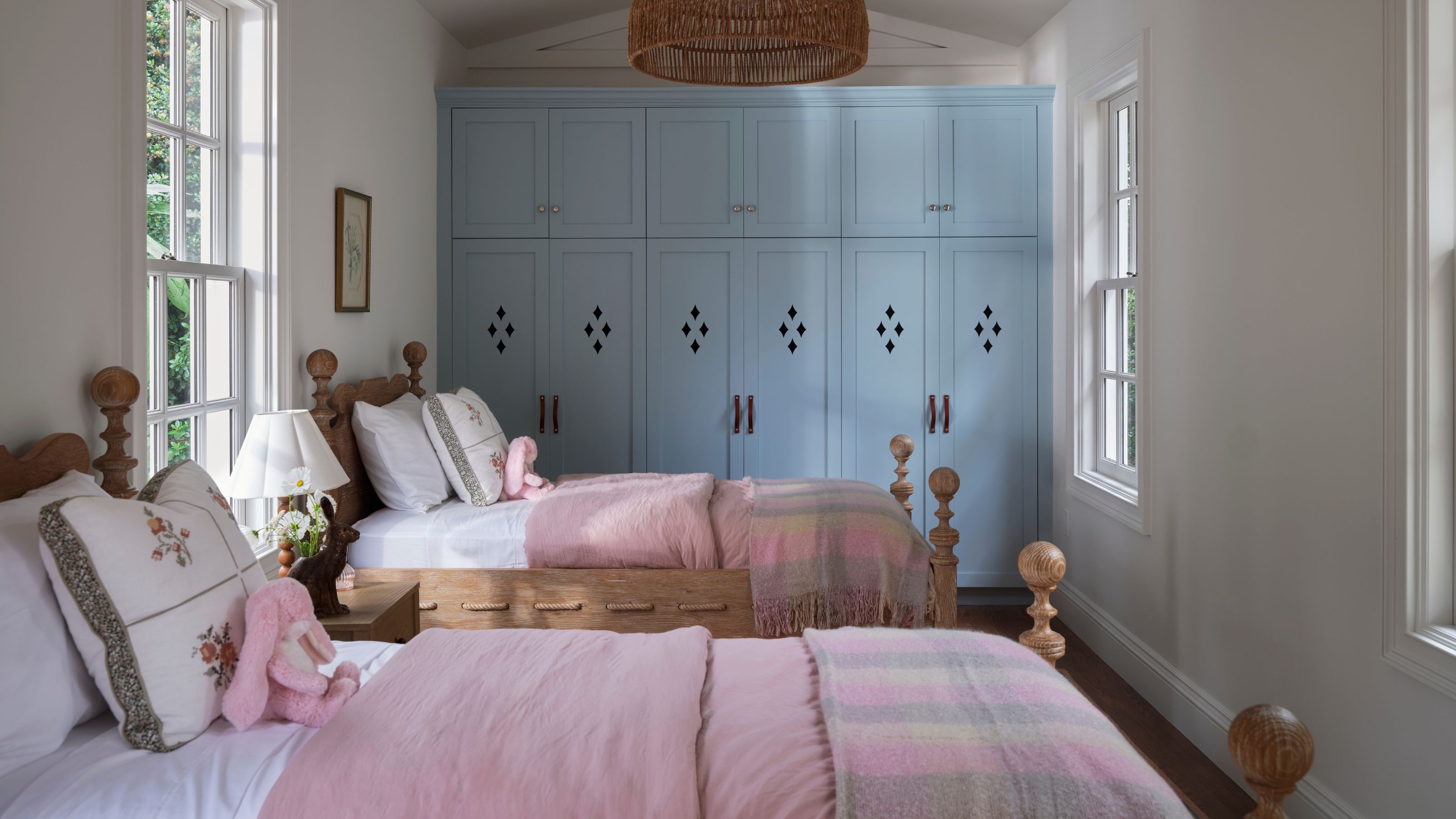 a white painted twin kids room with blue custom cabinetry, two antique pin beds styled with white and pink linens with floral pillows and small bunny toys