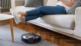 A robot vacuum cleaning a rug on hard floor while a woman sits on the couch with her feet up