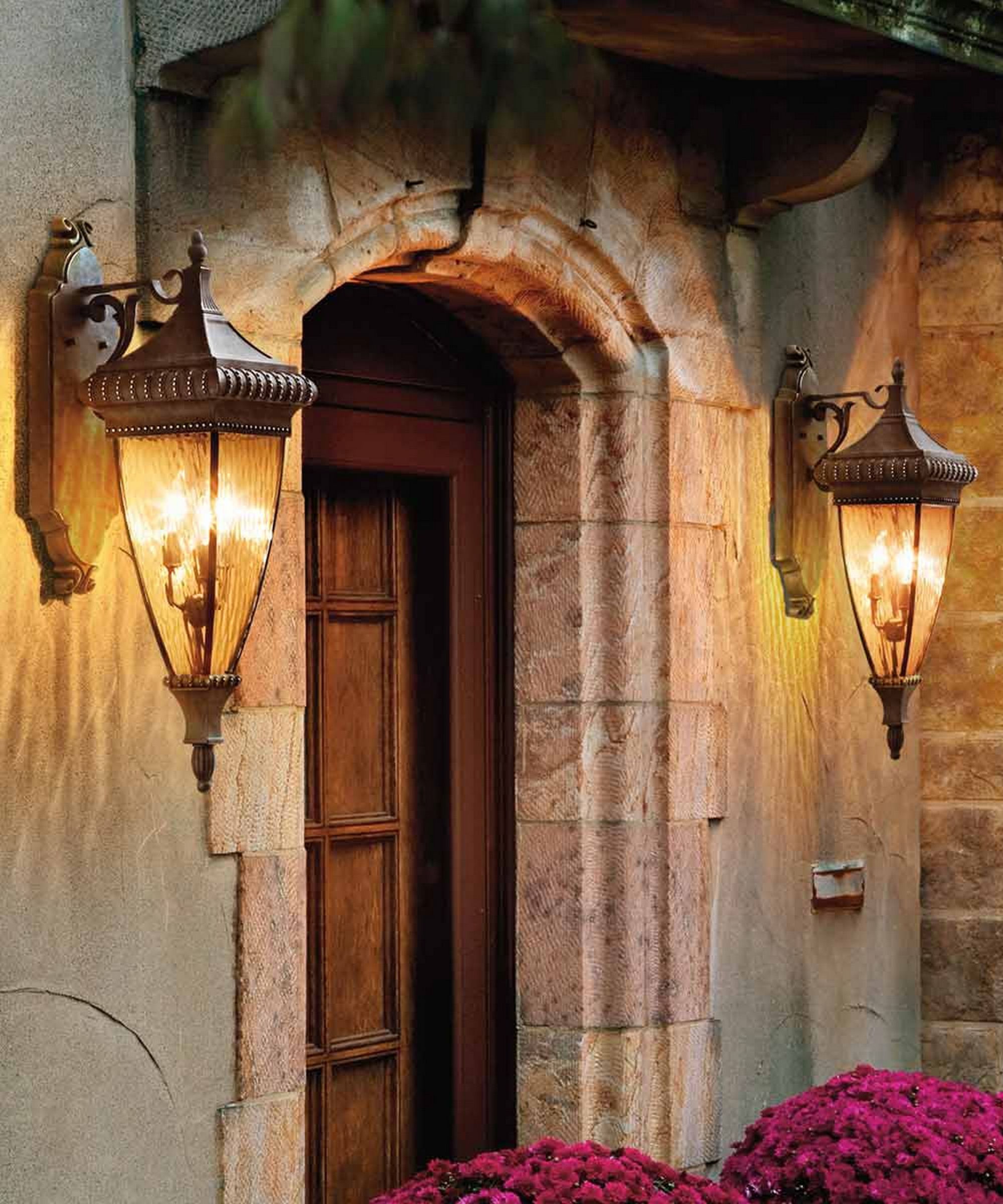 tone cottage porch with a wooden door framed by a rustic arch, lit by two ornate lantern-style wall lights, and flanked by vibrant pink flowers.