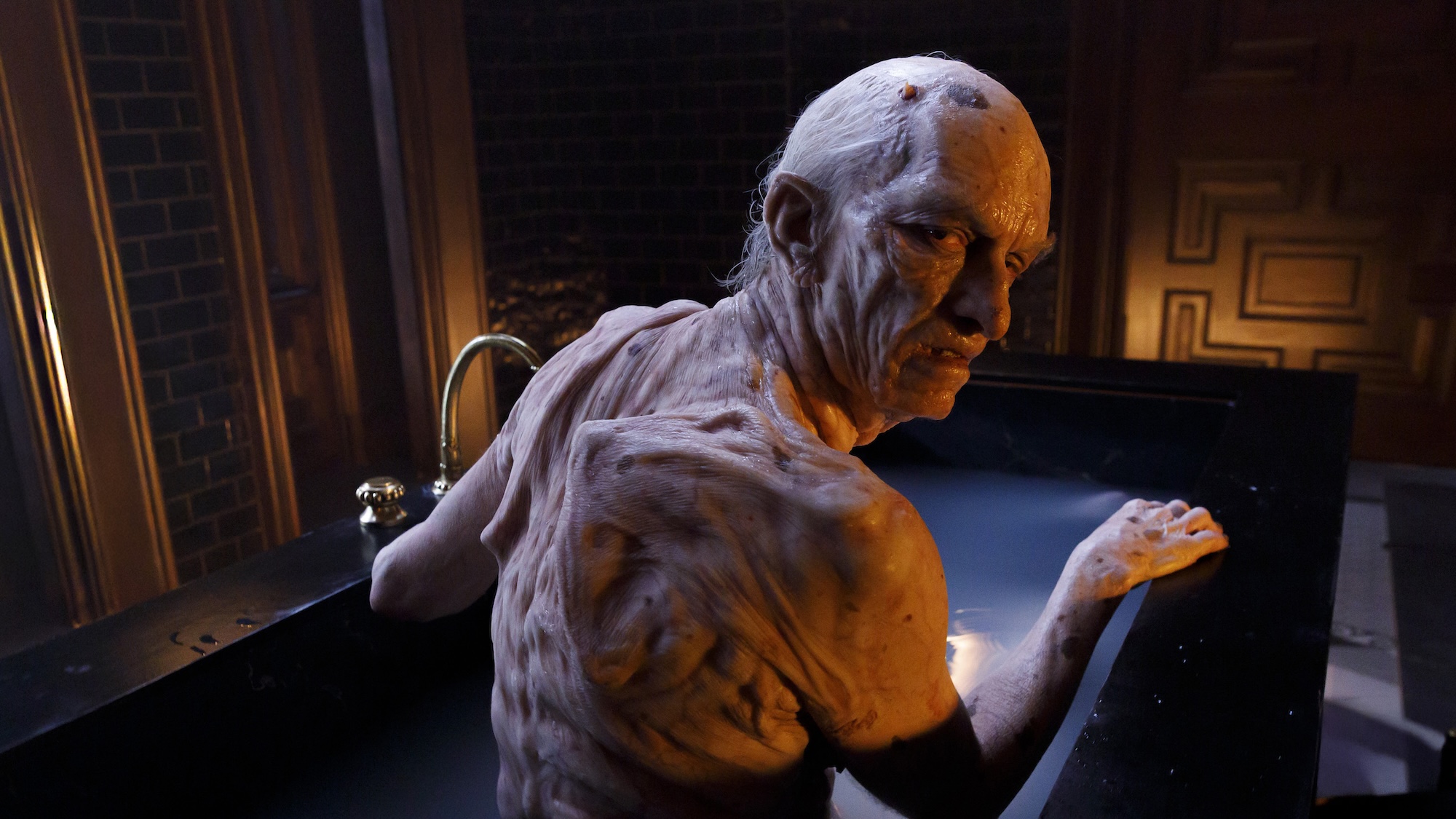 Mr. Nas nude with many wrinkles and bones protruding along his shoulder blades, sitting inside a black bathtub in the Penthouse in American Horror Story 13