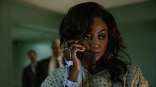 Raquel 'Raq' Thomas (Patina Miller) on the phone wearing a hospital gown and a cannula in her hand. Two detectives stand behind her, blurred into the background.