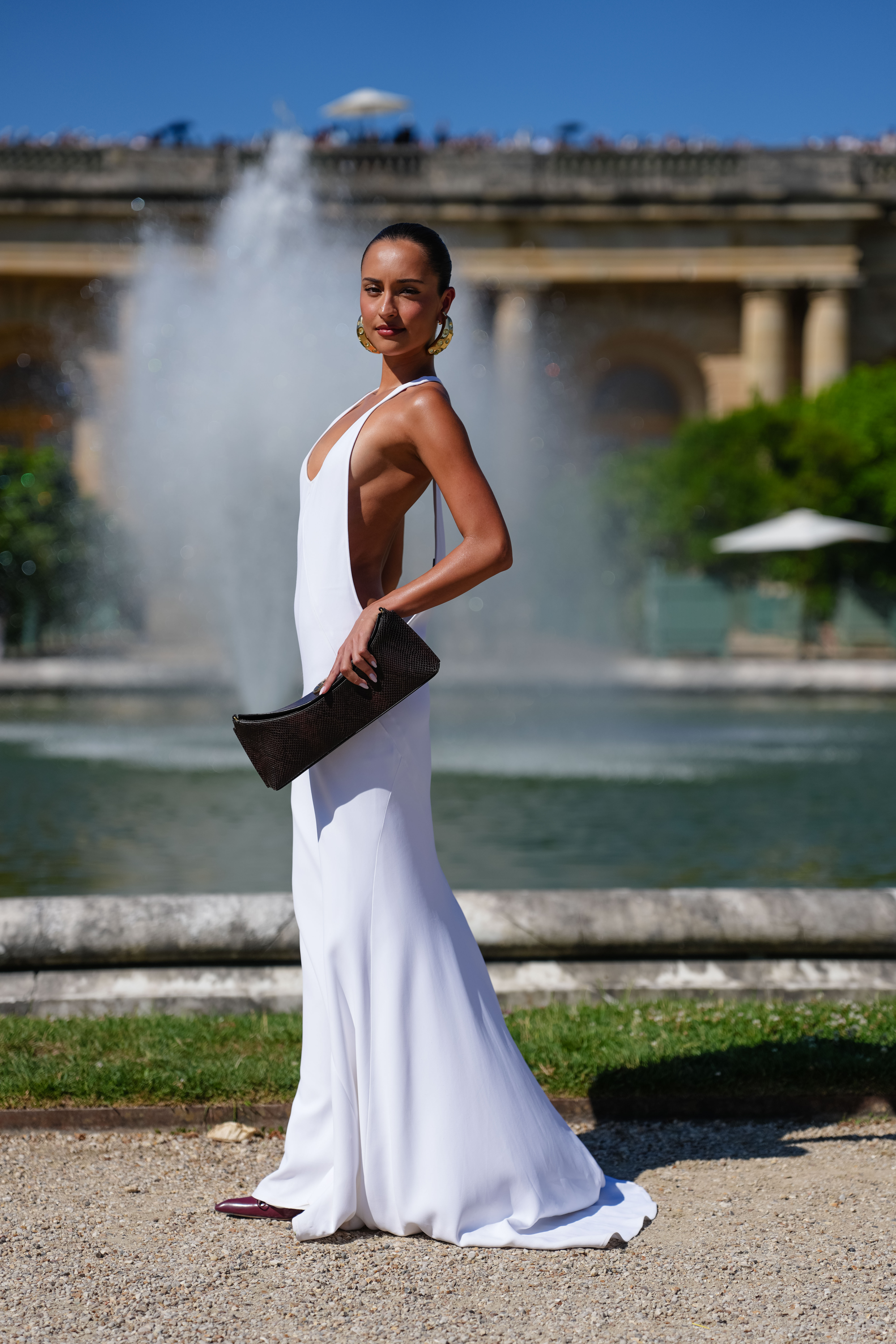 VERSAILLES, FRANCE - JUNE 29: Alexandra Saint Mleux wears a white floor-length halterneck dress with a plunging neckline, deep side cutouts and a flared train hem, styled with gold sculptural hoop earrings and a black textured leather clutch bag. Hair is pulled back into a sleek bun, outside Jacquemus, during the Paris Fashion Week - Menswear Spring/Summer 2026, on June 29, 2025 in Versailles, France.